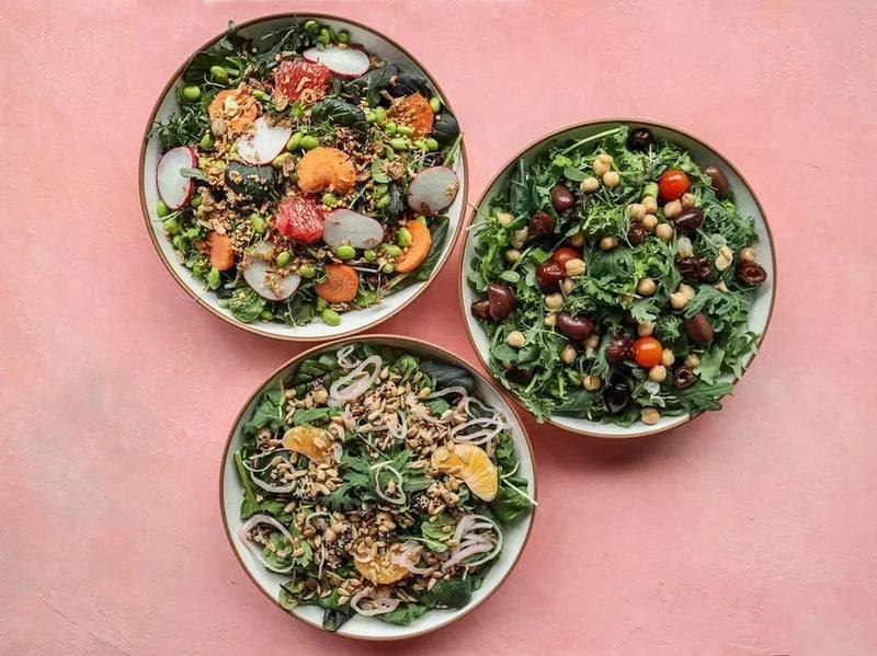 Three colorful fresh salads topped with chickpeas, citrus, seeds, and mixed vegetables on a wooden table.