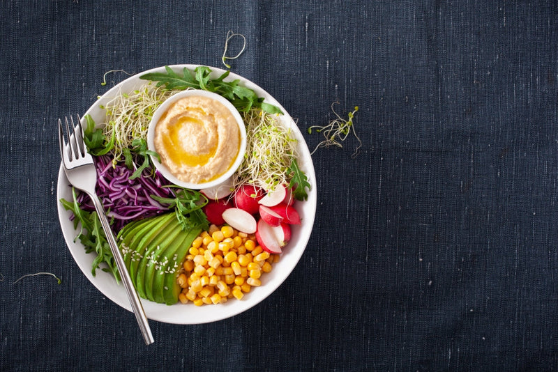  A vegan lunch bowl with sliced avocado, sweet corn, radishes, shredded purple cabbage, arugula, alfalfa sprouts, and a small bowl of hummus drizzled with olive oil, served on a white plate with a fork on a dark fabric background.